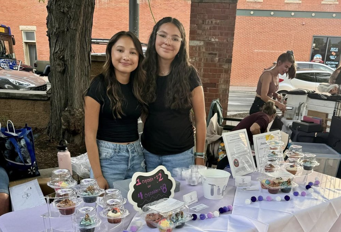 Two girls stand at a table and sell cupcakes.